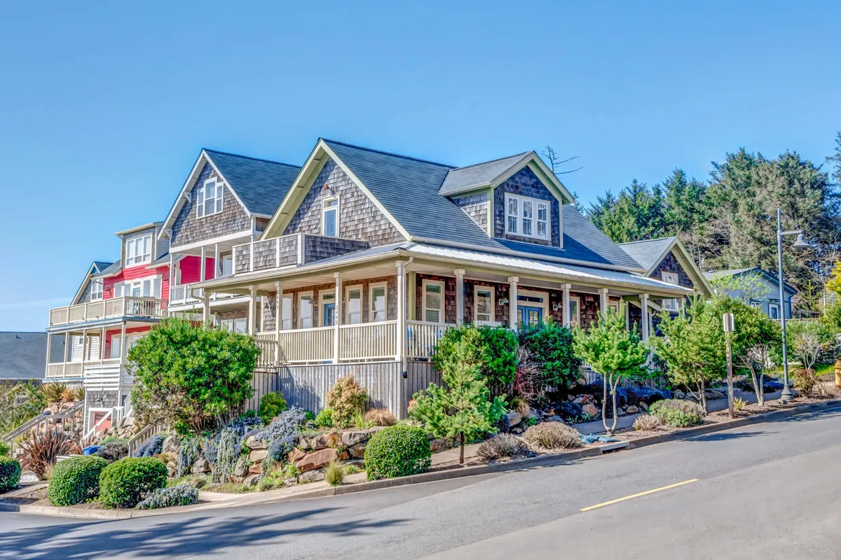The image showcases the inviting exterior of The Black Pearl, a charming two-level house with a distinctive wrap-around porch surrounded by lush landscaping. The scene is tranquil, evoking a sense of serenity, and highlights the architectural uniqueness of the home set within the upscale Olivia Beach community. The porch is perfectly positioned to enjoy sunset views or savor a morning coffee, while the garden adds to the home's relaxing ambiance.