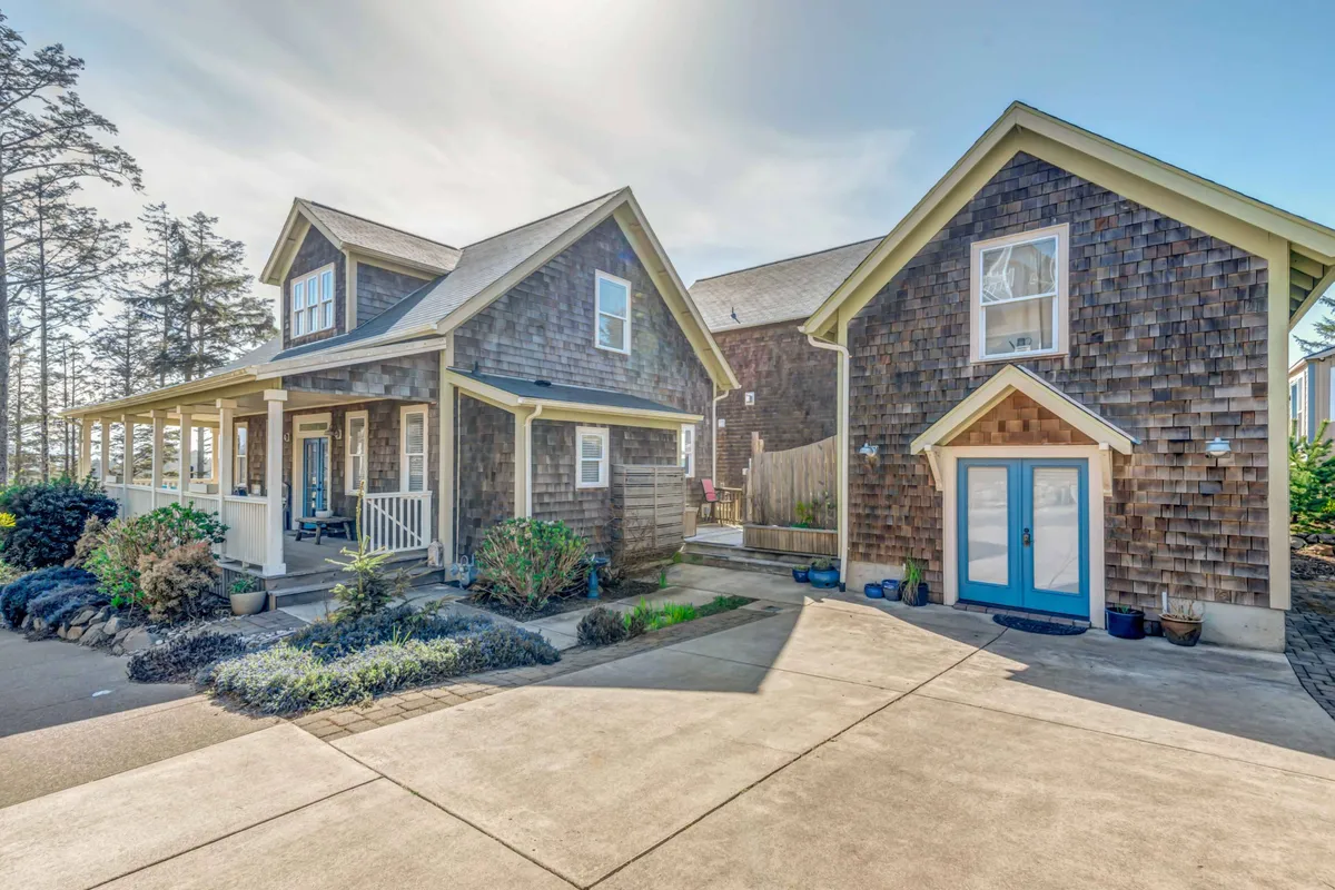 An exterior view of The Black Pearl, a two-story home with a welcoming blue front door, surrounded by a landscaped garden. The scene feels inviting and sets the stage for a family’s coastal getaway, connecting the home to the outdoors.