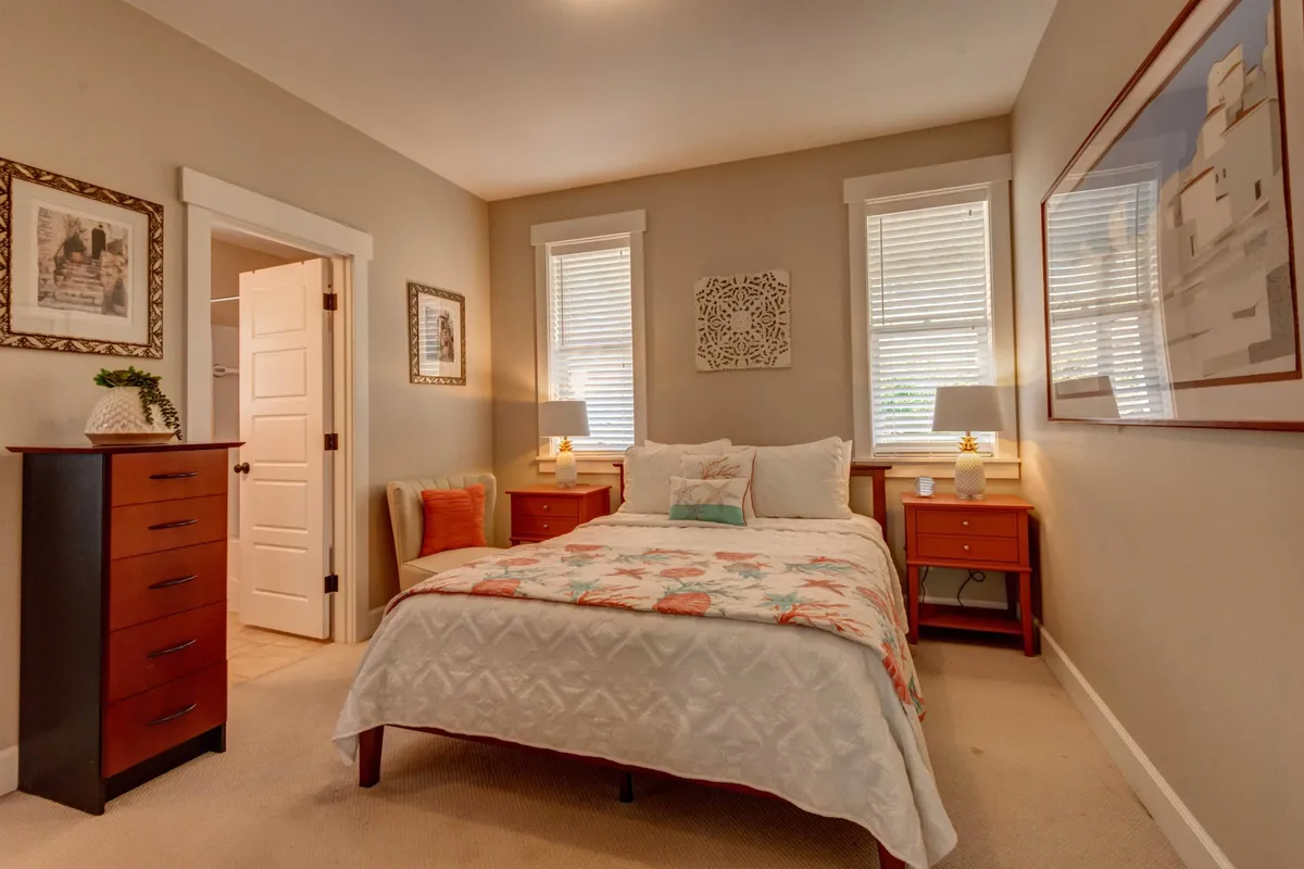A cozy bedroom featuring a bed with a floral quilt, matching nightstands with lamps, artwork on the walls, and a dresser. Soft natural light filters in, creating an inviting retreat perfect for resting and unwinding.