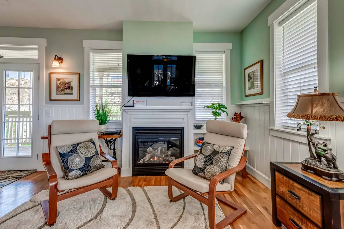 A sunlit living room featuring plush armchairs, a cozy gas fireplace at the center, a Smart TV, and warm decorative accents. The space is bathed in natural light from large windows, inviting relaxation and social gatherings.