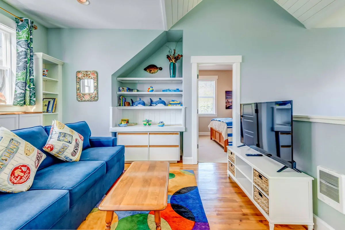 A vibrant media/game room with a blue couch, colorful rug, shelves stocked with decorative items and games, and a TV unit. The atmosphere is lively yet cozy—perfect for relaxing, playing games, or movie nights with family.
