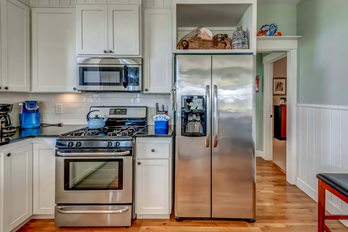 This image showcases a warm and welcoming kitchen with a stainless steel refrigerator, gas stove, blue kettle, and crisp white cabinetry. The space feels inviting and perfectly suited for group cooking and shared meals, with plenty of room for gathering around.