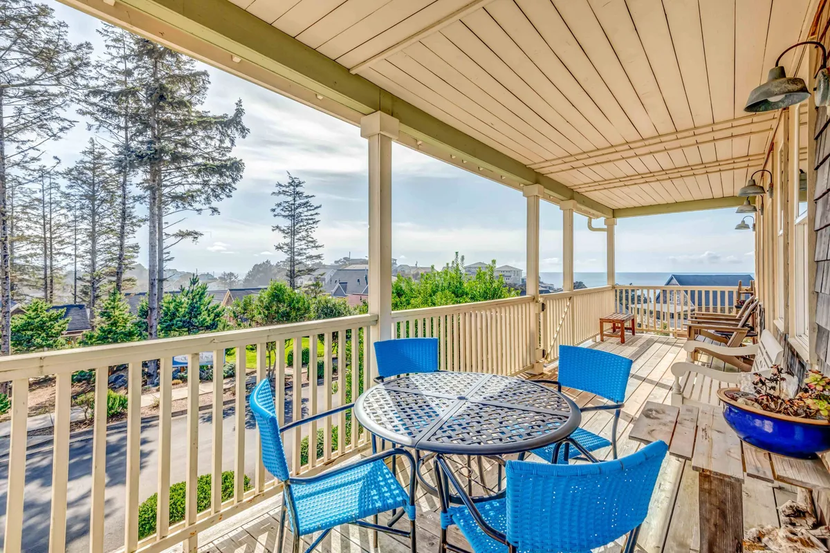A cheerful outdoor dining area with blue chairs and a round table sits on the deck, surrounded by views of trees and the ocean. The space invites guests to dine alfresco or gather for barbecues while enjoying coastal breezes and vibrant sunsets.