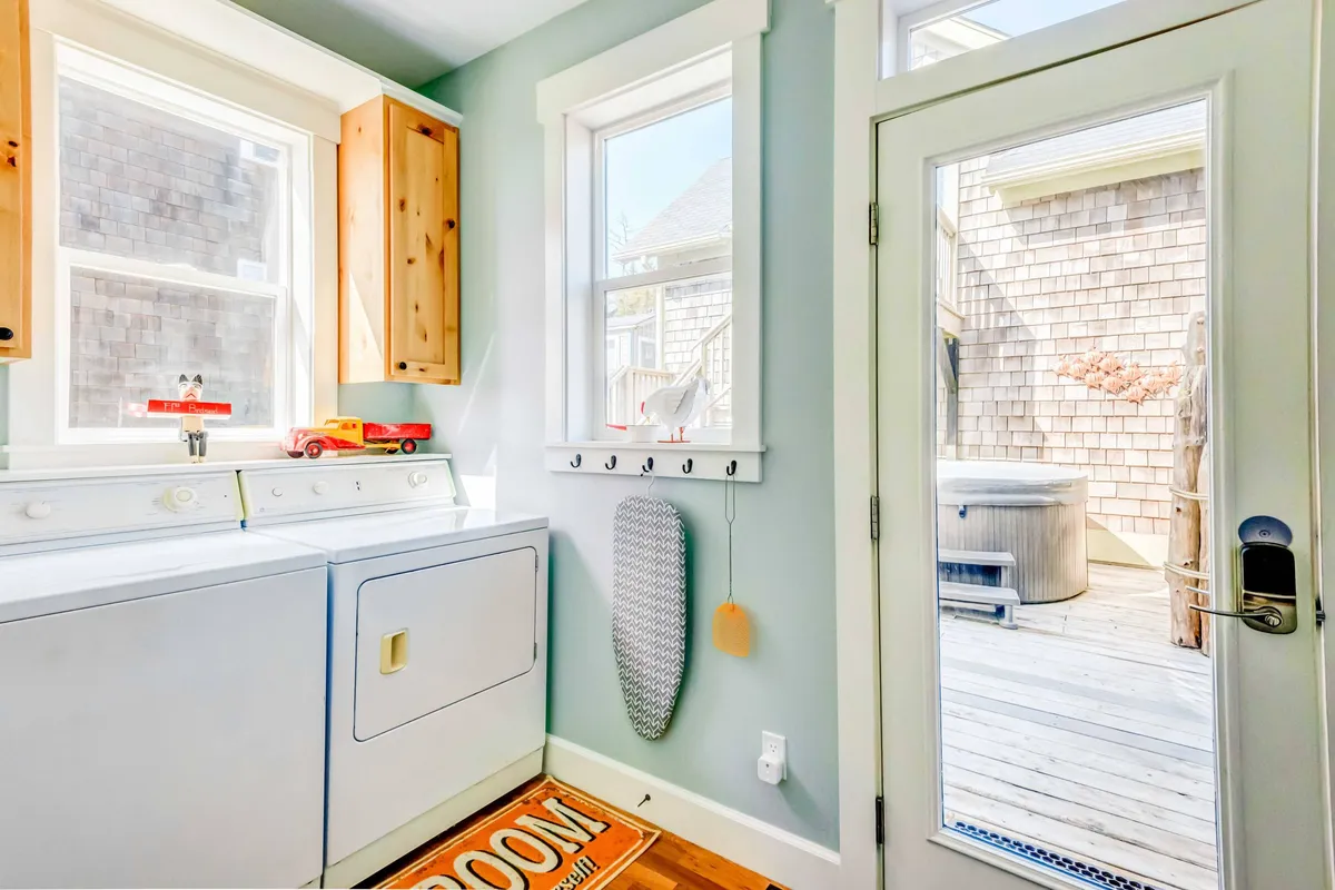 This image features a functional laundry space with a washing machine, dryer, iron board, and a door that leads out to a deck where a jacuzzi is visible. The connection between the indoor utility area and the outdoor relaxation space is highlighted.