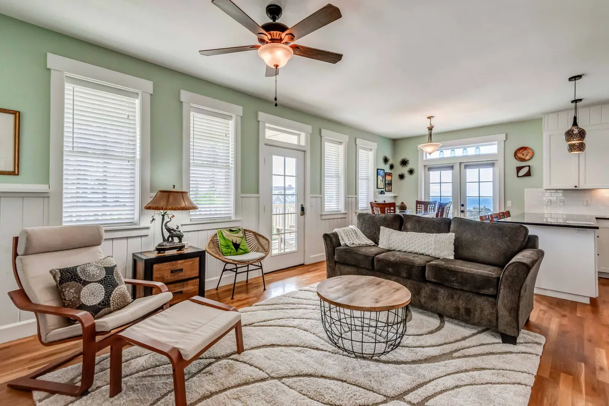 A cozy living room with a comfortable sofa and armchair is bathed in natural light from large windows. The setup offers a welcoming retreat after a day spent walking the beach or playing fetch, and the view through the windows hints at the home's proximity to the scenic outdoors.