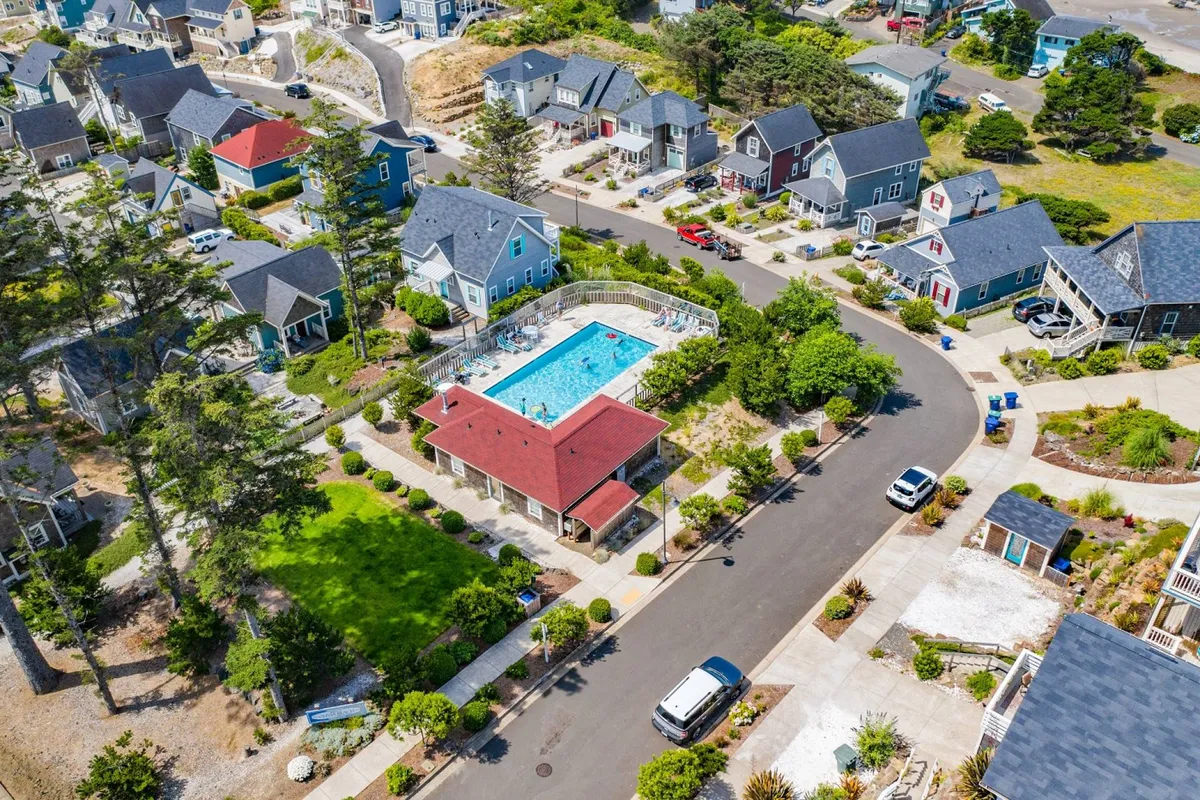 An exterior view of the Olivia Beach community showing the resort-style swimming pool at the center, surrounded by green lawns, the community center, and neighboring houses. The space is lively, bathed in sunlight, and ready for swimming, sunbathing, and socializing.