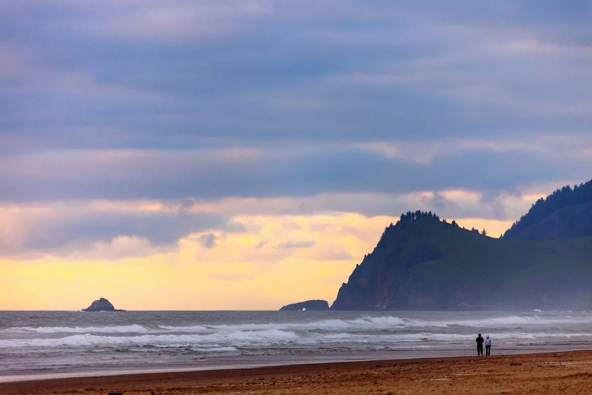 This image captures a couple walking along a sandy beach, with distant cliffs and waves crashing on the shore. The scene is bathed in warm sunset light, evoking a sense of tranquility and romance. The expanse of ocean and dramatic coastline invite exploration and wildlife watching, perfectly illustrating the coastal adventure described in the section.